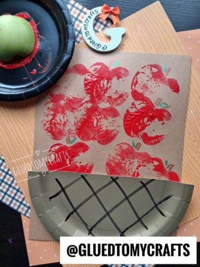 A craft project shows red apples stamped on paper, made using half an apple and red paint. Theres a paper plate designed to look like a basket and a @GLUEDTOMYCRAFTS label at the bottom.