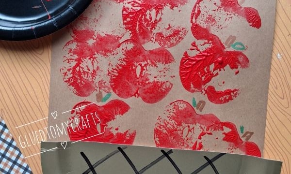 A craft project shows red apples stamped on paper, made using half an apple and red paint. Theres a paper plate designed to look like a basket and a @GLUEDTOMYCRAFTS label at the bottom.