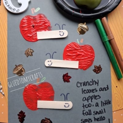 Gray paper with red apple prints, craft sticks as snail bodies, and drawn smiles. Fall leaf stickers and handwritten note: Crunchy leaves and apples too; a little fall snail says hello to you! Scattered markers and an apple nearby.