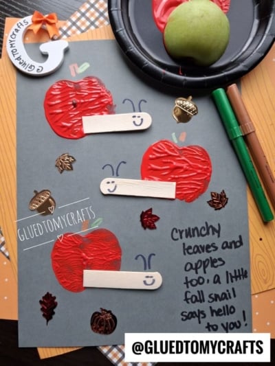 Gray paper with red apple prints, craft sticks as snail bodies, and drawn smiles. Fall leaf stickers and handwritten note: Crunchy leaves and apples too; a little fall snail says hello to you! Scattered markers and an apple nearby.