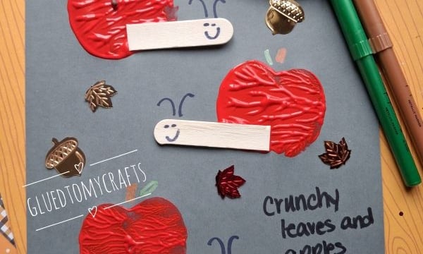 Gray paper with red apple prints, craft sticks as snail bodies, and drawn smiles. Fall leaf stickers and handwritten note: Crunchy leaves and apples too; a little fall snail says hello to you! Scattered markers and an apple nearby.