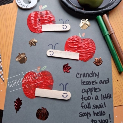 Gray paper with three red apple crafts made of paint and craft sticks, decorated with smiling faces. Surrounded by fall-themed embellishments, markers, an apple, and a paint plate. Handwritten note reads a message about snails and apples.