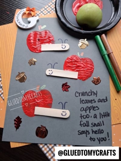 Gray paper with three red apple crafts made of paint and craft sticks, decorated with smiling faces. Surrounded by fall-themed embellishments, markers, an apple, and a paint plate. Handwritten note reads a message about snails and apples.