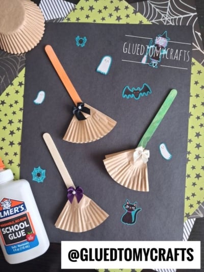 Halloween craft scene featuring a cupcake liner broom, with popsicle stick witch brooms crafted from colorful cupcake liners and decorated with small bows. The scene is surrounded by Halloween stickers, a bottle of school glue, and set against a black background. GLUEDTOMYCRAFTS label is visible.