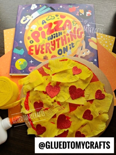 Children’s book A Pizza with Everything On It is pictured alongside a cardboard pizza craft made from tissue paper and glitter heart shapes, surrounded by crafting supplies and orange paper. @GLUETOMYCRAFTS is prominently displayed.