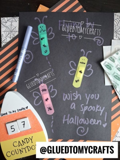 A black paper decorated with colorful Band-Aid Butterfly Craft butterflies, whimsical white doodles, and “Wish you a spooky Halloween!” is surrounded by craft supplies and a candy countdown card. The handle @GLUEDTOMYCRAFTS is displayed.