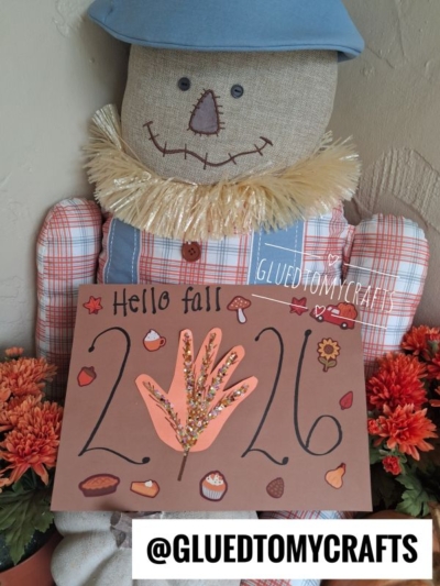 A scarecrow doll holds a brown Hello Fall 2023 craft sign decorated with leaves and pumpkins, surrounded by orange flowers—making it a perfect inspiration for FALL CRAFTS FOR KIDS. The Instagram handle @GLUETOMYCRAFTS is shown at the bottom.