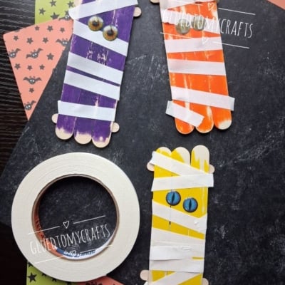 Four popsicle sticks crafts painted as mummies with googly eyes, wrapped in white tape, on Halloween-themed paper. A roll of tape and the Instagram handle @GLUEDTOMYCRAFTS are also visible.