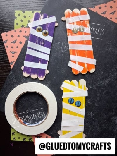Four popsicle sticks crafts painted as mummies with googly eyes, wrapped in white tape, on Halloween-themed paper. A roll of tape and the Instagram handle @GLUEDTOMYCRAFTS are also visible.