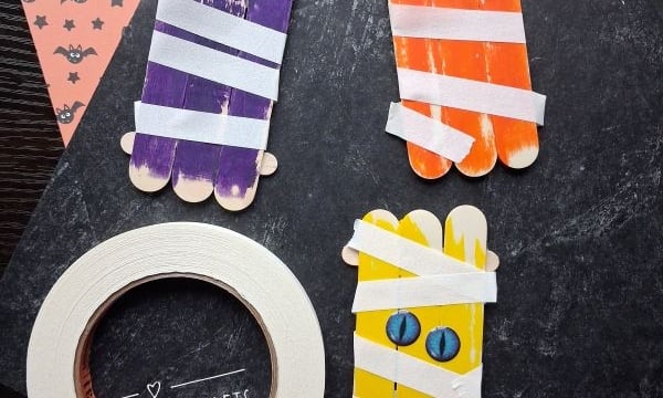 Four popsicle sticks crafts painted as mummies with googly eyes, wrapped in white tape, on Halloween-themed paper. A roll of tape and the Instagram handle @GLUEDTOMYCRAFTS are also visible.