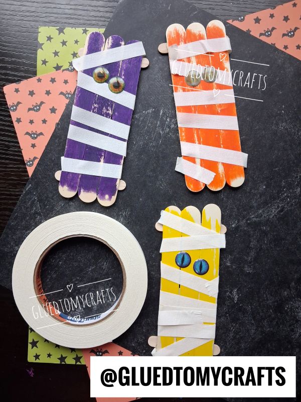 Four popsicle sticks crafts painted as mummies with googly eyes, wrapped in white tape, on Halloween-themed paper. A roll of tape and the Instagram handle @GLUEDTOMYCRAFTS are also visible.
