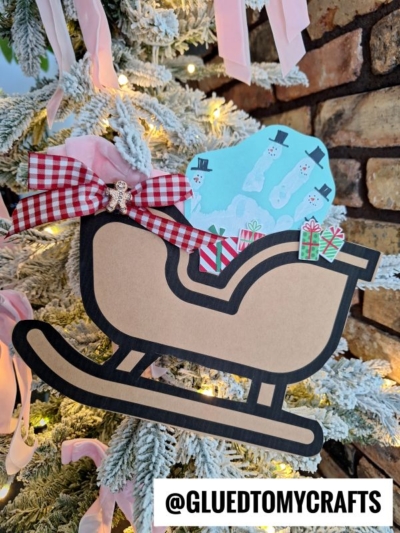 A charming Christmas sleigh keepsake ornament with a red checked bow, small snowman card, and tiny gifts hangs on a snowy Christmas tree. A brick wall is in the background. Text reads @GLUEDTOMYCRAFTS.
