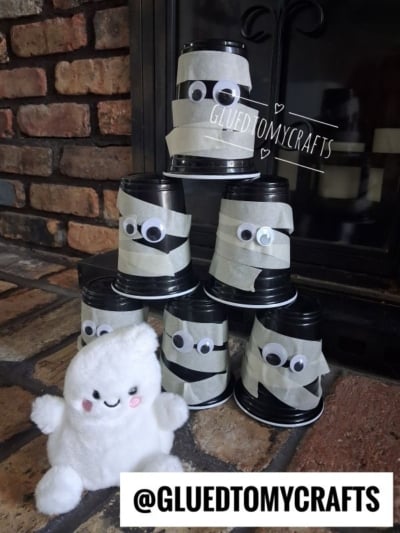 Six black cups decorated as mummies with googly eyes are stacked in a pyramid, making for fun Halloween crafts for kids. A fluffy white smiling plush toy sits in front, with a brick fireplace in the background. @GLUEDTOMYCRAFTS is written at the bottom.