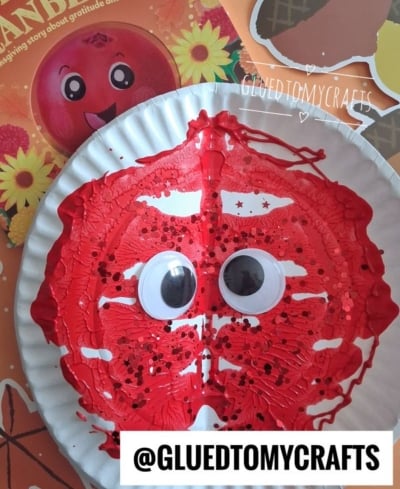 A paper plate painted red with black dots and two large googly eyes, resembling a cranberry, placed on a colorful background. Text @GLUEDTOMYCRAFTS is displayed at the bottom.