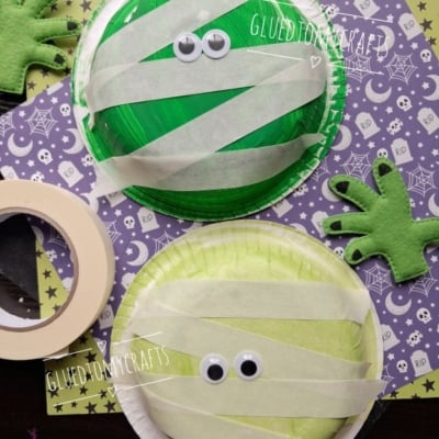 Two paper plate mummy crafts with googly eyes and tape strips on green and yellow plates, surrounded by felt monster hands, purple Halloween-themed paper, and a roll of masking tape. @GLUEDTOMYCRAFTS is visible.