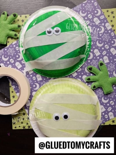 Two paper plate mummy crafts with googly eyes and tape strips on green and yellow plates, surrounded by felt monster hands, purple Halloween-themed paper, and a roll of masking tape. @GLUEDTOMYCRAFTS is visible.