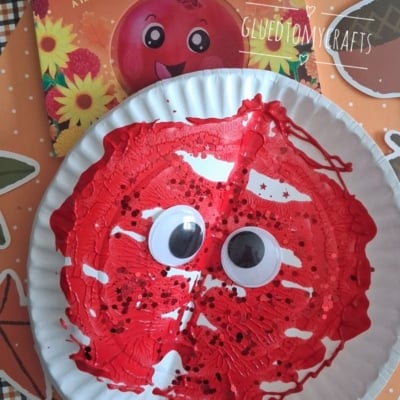 A paper plate painted red with large googly eyes and black sprinkles, resembling a cranberry, sits in front of a cranberry-themed childrens book on a festive autumn-patterned surface.