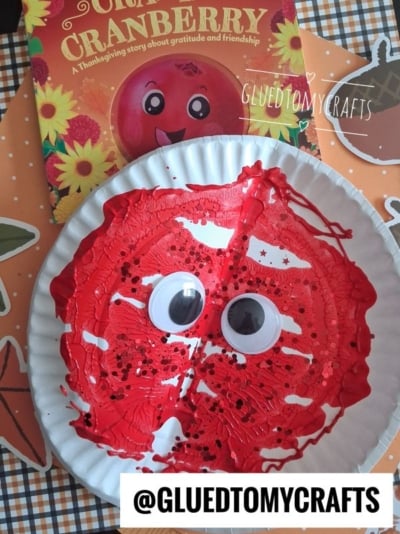 A paper plate painted red with large googly eyes and black sprinkles, resembling a cranberry, sits in front of a cranberry-themed childrens book on a festive autumn-patterned surface.