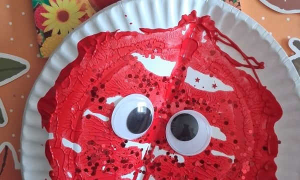 A paper plate painted red with large googly eyes and black sprinkles, resembling a cranberry, sits in front of a cranberry-themed childrens book on a festive autumn-patterned surface.