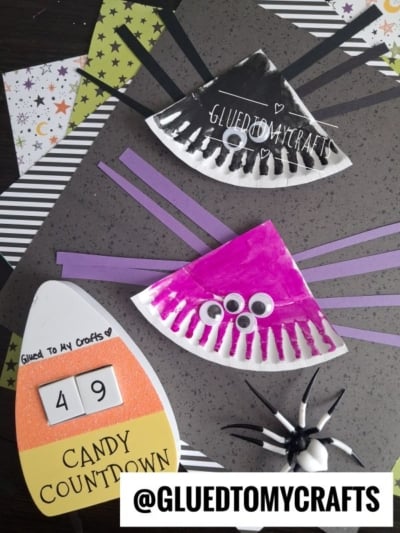 Colorful Halloween crafts for kids: black-and-purple paper plate spiders with googly eyes and yarn legs, a candy corn countdown sign showing 49 days, and a plastic spider on a dark table. @GLUETOMYCRAFTS watermark is visible.