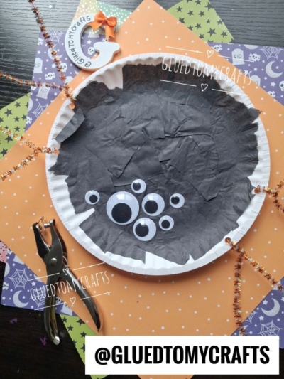 A paper plate spider craft featuring black tissue paper, googly eyes, and orange sparkly pipe cleaners for legs sits atop festive Halloween paper—perfect for Halloween Crafts for Kids. Hole punch and @GLUEDTOMYCRAFTS sign are included.