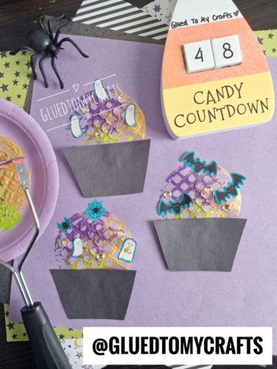 Purple paper displays three charming Halloween cupcake crafts, each decorated with ghost and bat stickers—perfect for Halloween Crafts for Kids. Nearby are a spider, waffle stamp, and a candy countdown sign. @GLUEDTOMYCRAFTS is written on the bottom.
