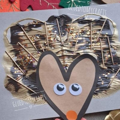 A handmade hedgehog craft using googly eyes, paper, toothpicks, paint, and glitter is shown on a gray background with autumn leaf cutouts. @GLUEDTOMYCRAFTS is labeled at the bottom.