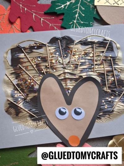 A handmade hedgehog craft using googly eyes, paper, toothpicks, paint, and glitter is shown on a gray background with autumn leaf cutouts. @GLUEDTOMYCRAFTS is labeled at the bottom.