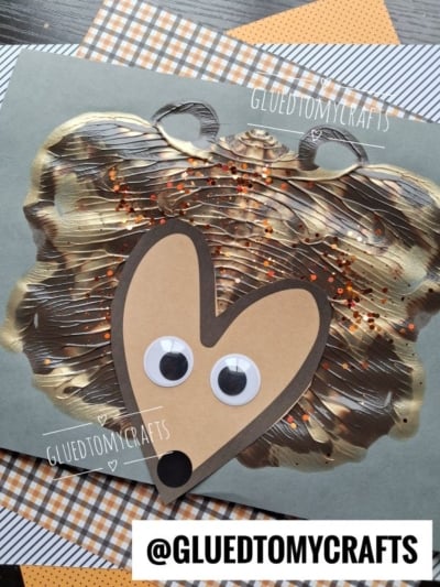 A paint splat hedgehog craft made with brown paint, orange glitter, and googly eyes on paper. The image features the @GLUETOMYCRAFTS watermark on a plaid-patterned background.