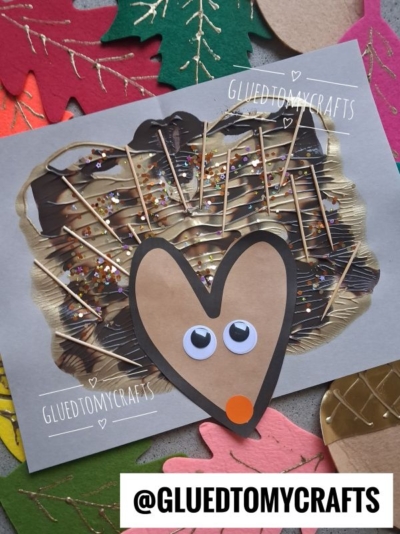 A handmade hedgehog craft with a heart-shaped face, googly eyes, orange nose, and toothpick spines on a painted background. Surrounded by colorful paper leaves.