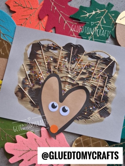 A handmade hedgehog craft with a brown paper face, googly eyes, and decorated with glitter on a painted background, surrounded by colorful paper leaves. The handle @GLUEDTOMYCRAFTS is visible.