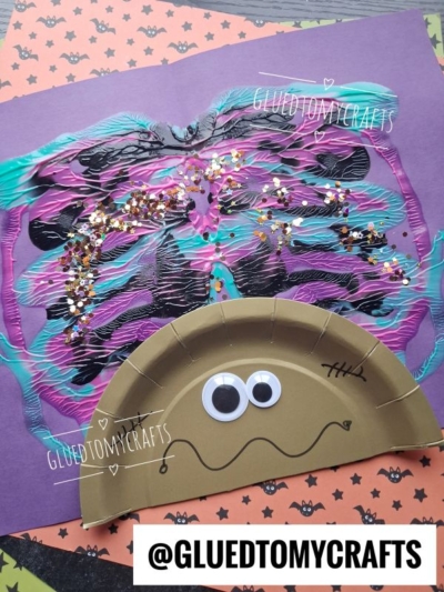A paper plate spider with googly eyes is attached to a colorful, painted web with glitter on purple paper. Halloween-themed papers are in the background. Text reads @GLUEDTOMYCRAFTS.