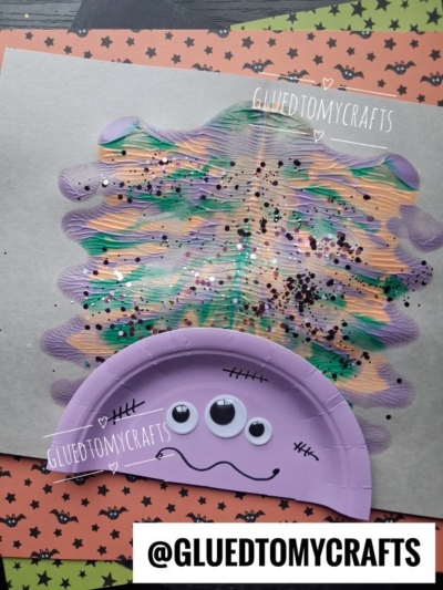 A Halloween craft features a purple paper plate monster with googly eyes and marker details, topped with a painted, glittery, colorful hair section on gray paper. The background includes orange and green star-patterned paper.