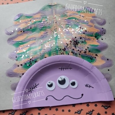 A purple paper plate monster with three googly eyes and black stitches is glued to a painted, glittery background. The craft is on Halloween-themed paper. @GLUEDTOMYCRAFTS is labeled on the image.