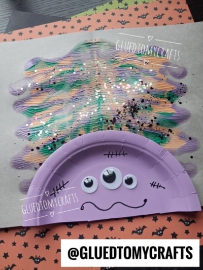 A purple paper plate monster with three googly eyes and black stitches is glued to a painted, glittery background. The craft is on Halloween-themed paper. @GLUEDTOMYCRAFTS is labeled on the image.