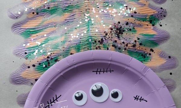 A purple paper plate monster with three googly eyes and black stitches is glued to a painted, glittery background. The craft is on Halloween-themed paper. @GLUEDTOMYCRAFTS is labeled on the image.