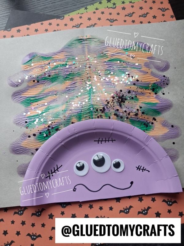 A purple paper plate monster with three googly eyes and black stitches is glued to a painted, glittery background. The craft is on Halloween-themed paper. @GLUEDTOMYCRAFTS is labeled on the image.