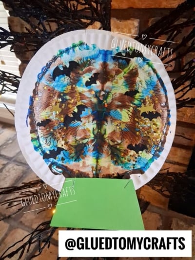 A colorful, symmetrical butterfly made with paint on a paper plate—a perfect example of PAINT SPLAT CRAFTS—displayed in front of a brick background. The text @GLUEDTOMYCRAFTS appears in the image.