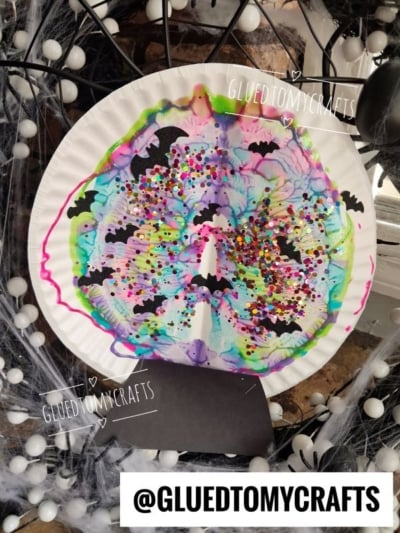 Colorful paper plate craft with marker tie-dye, black bat shapes, confetti, and a web background—perfect for fans of PAINT SPLAT CRAFTS. @GLUEDTOMYCRAFTS is written on the image in several places.