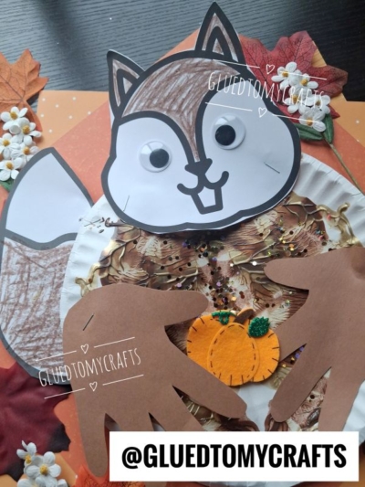 A squirrel craft made from a paper plate, googly eyes, brown paper hand cutouts for arms, and a small pumpkin. Surrounded by fake leaves and flowers on a polka dot background.