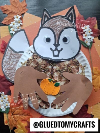 A squirrel craft made from a paper plate, colored paper, and googly eyes holds a small orange pumpkin, surrounded by fake autumn leaves and white flowers. @GLUEDTOMYCRAFTS is written at the bottom.