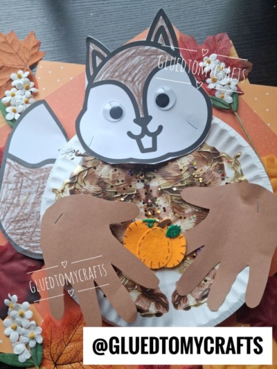 A squirrel craft made from paper, googly eyes, two handprint cutouts, and leaves, holding a small pumpkin decoration. Text reads @GLUEDTOMYCRAFTS..