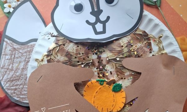 A squirrel craft made from paper, googly eyes, two handprint cutouts, and leaves, holding a small pumpkin decoration. Text reads @GLUEDTOMYCRAFTS..