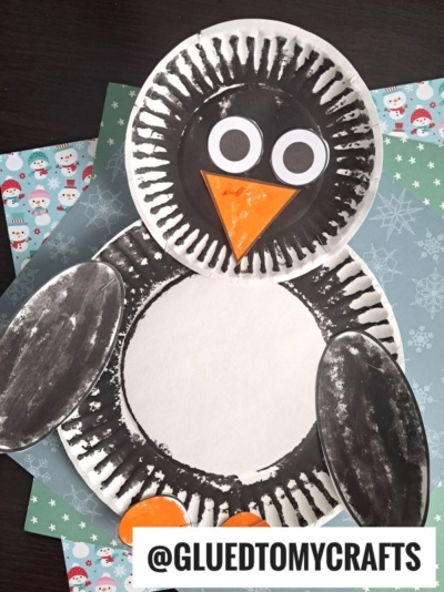 A paper plate penguin craft made from painted paper plates, construction paper eyes and beak, displayed on winter-themed paper. @GLUEDTOMYCRAFTS text is at the bottom. This charming paper penguin project is perfect for celebrating the snowy season!.