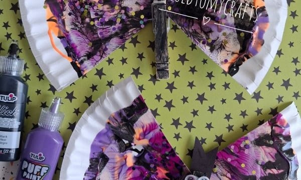 Two colorful butterfly crafts made from paper plates and paint, with decorated clothespins for bodies and googly eyes, are displayed on a patterned surface with puffy paint bottles nearby. Text reads @GLUEDTOMYCRAFTS.