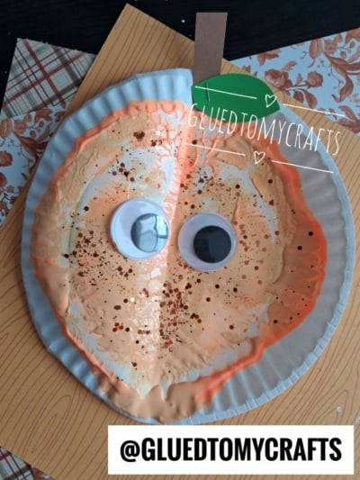 Painted paper plate pumpkin with two large googly eyes, brown stem, green leaf, and orange sparkles, displayed on patterned paper backgrounds. GLUEDTOMYCRAFTS watermark and tag are visible.