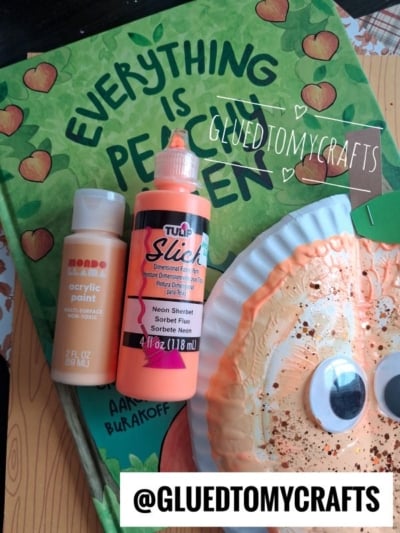 A bottle of acrylic paint and neon fabric paint sit on the book Everything is Peachy Keen, next to a paper peach craft with a face. @GLUEDTOMYCRAFTS is written at the bottom.