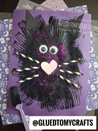 A fork painted cat craft made with black paint, paper, googly eyes, straws for whiskers, and a pink heart nose on purple paper, with Halloween-themed papers in the background.