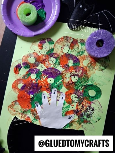 A Halloween craft features a handprint on paper, surrounded by colorful paint circles made with foam donuts in green, orange, purple, and black. A plate and foam donuts sit nearby, and a bat decoration is visible.