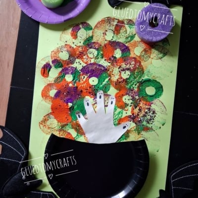 Childrens Halloween craft: Colorful circular sponge prints make bubbles from a black paper cauldron with a white handprint on green paper. Bat decorations and sponges are nearby. Text reads @GLUEDTOMYCRAFTS..